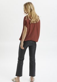Blouse couleur rouille à manches courtes, associée à un pantalon en cuir noir évasé et des sandales de couleur claire, vue de dos.