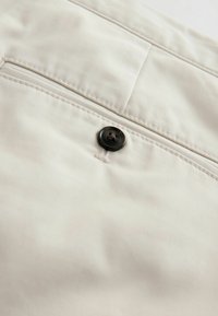 Tissu beige avec un bouton foncé cousu près d'une couture horizontale et un petit détail de poche au-dessus du bouton.