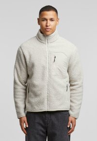 Fleece-hvid jakke med høj krave, frontlommer med lynlås og tekstureret stof. Har en venstre brystlomme med lynlåslukning.