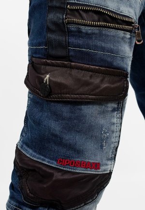 Denim jeans in het blauw en zwart, met een ritszak, leren accenten en rode logo stiksels. Voorzien van een verouderde textuur met meerdere panelen.