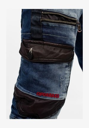 Denim jeans in het blauw en zwart, met een ritszak, leren accenten en rode logo stiksels. Voorzien van een verouderde textuur met meerdere panelen.