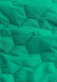 Tissu matelassé vert avec un motif hexagonal, présentant des détails de couture visibles. La surface a une texture lisse, légèrement froissée.