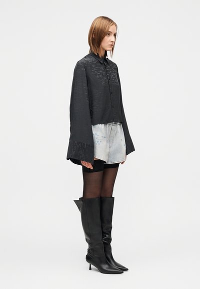 Μαύρο κοντό πουκάμισο με υφή, ψηλό γιακά και φαρδιά μανίκια, συνδυασμένο με σορτς από τζιν και μαύρες δερμάτινες μπότες μέχρι το γόνατο.