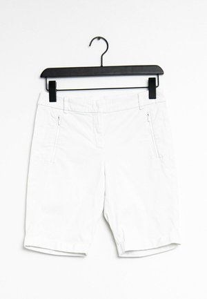 Pantalones cortos blancos de tela ligera, con dos bolsillos laterales con cremallera, puños enrollados y una textura suave. Colgados en una percha negra de clip.