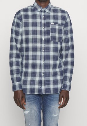 Personne portant une chemise à carreaux bleue et blanche à boutons avec une poche poitrine, associée à un jean bleu clair déchiré.