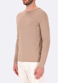 Camicia beige a maniche lunghe realizzata in tessuto morbido, con collo rotondo e silhouette aderente, indossata con pantaloni bianchi, dettagli di design semplici.