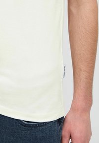 Tricou din bumbac galben deschis, cu etichetă pe lateral, decolteu rotund și mâneci scurte, purtat împreună cu blugi din denim închis.
