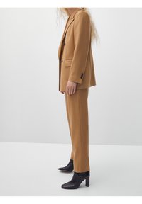 En person står i profil, klädd i en stilren kamelfärgad kostym med en skräddarsydd kavaj och matchande byxor, kompletterad med eleganta svarta ankelboots.