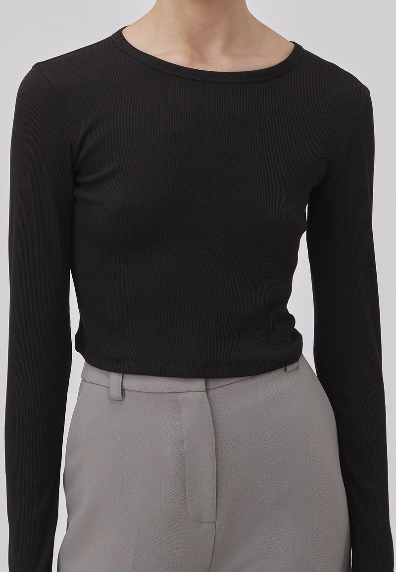 Personne portant un haut cintré noir à manches longues côtelé, rentré dans un pantalon taille haute gris clair, sur mesure, avec des passants pour ceinture.