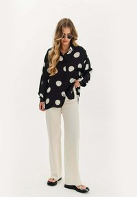 Czarna koszula zapinana na guziki z dużymi białymi kropkami, w połączeniu z kremowymi spodniami o szerokich nogawkach i czarnymi płaskimi sandałami. Modelka nosi okulary przeciwsłoneczne oraz naszyjniki.