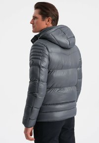 Giacca imbottita grigia con cappuccio; presenta sezioni trapuntate, polsini elasticizzati e un logo dettagliato sulla manica. Tessuto liscio e lucido.