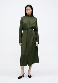 Largo vestido verde oliva con un alto cuello, detalle de botones, cintura ceñida con cinturón y mangas largas, hecho de una tela suave y fluida.