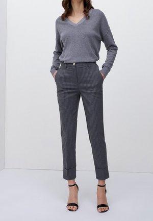 Grauer V-Ausschnitt-Pullover mit langen Ärmeln, kombiniert mit grauen Streifenhose mit umgeschlagenem Saum und vorderem Knopfverschluss. Schwarze Knöchelriemen.