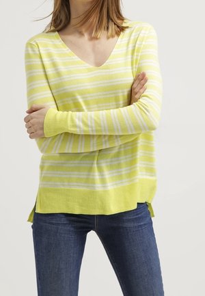 Žena nosící žluto-bílý proužkovaný svetr s výstřihem do V a dlouhými rukávy, v kombinaci s tmavě modrými džíny, se zkříženýma rukama na hrudi.