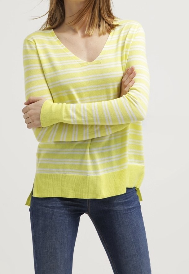 Femme portant un pull à col en V rayé jaune et blanc à manches longues, associé à un jean bleu foncé, les bras croisés sur la poitrine.