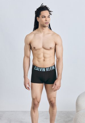 Μυώδης άνδρας με ντέντλοκς που φοράει μαύρα μποξεράκια Calvin Klein, στέκεται μπροστά σε απλό ανοιχτό φόντο, κοιτάζοντας προς το πλάι.