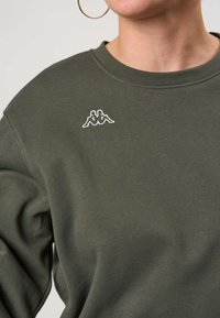 Persoană purtând un hanorac verde măsliniu cu un mic logo alb Kappa pe partea stângă a pieptului și o cerceafă mare vizibilă pe urechea stângă.