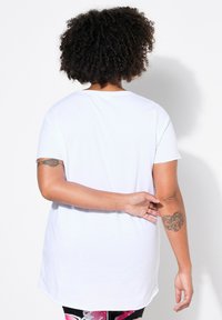 T-shirt blanc à manches courtes avec un col rond, présentant une coupe ample et un ourlet incurvé, vu de dos. Tatouage visible sur le bras gauche.