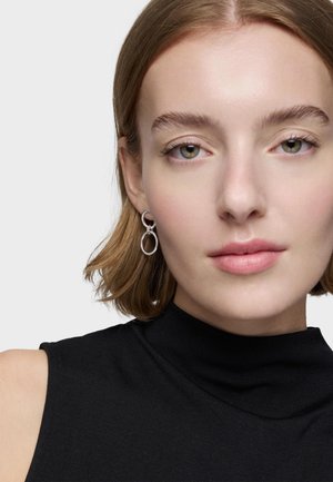 Kobieta o krótkich jasnobrązowych włosach, nosząca srebrne kolczyki koła i czarną, bezrękawnikową bluzkę z golfem, zwrócona przodem, o neutralnym wyrazie twarzy.