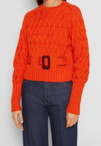 Pull en tricot orange avec un motif de losanges en relief, agrémenté d'une ceinture avec une boucle carrée à la taille, associé à un jean bleu foncé.