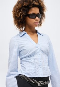 Γυναίκα με σγουρά μαλλιά που φοράει μαύρα γυαλιά ηλίου, φωτεινό γαλάζιο ριγέ πουκάμισο με κουμπιά και μαύρη ζώνη με καρφιά σε μαύρο παντελόνι.