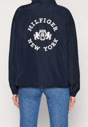 Personne portant une veste bleu marine avec un texte blanc "Hilfiger New York" et un logo blason dans le dos, assortie d'un jean bleu.