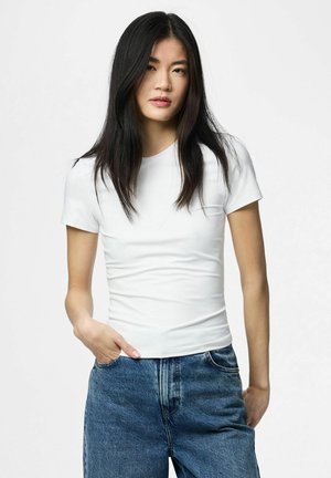 Junge Frau mit langen schwarzen Haaren, die ein eng anliegendes weißes Kurzarmshirt und lockere blaue Jeans trägt, eine Hand in der Tasche.