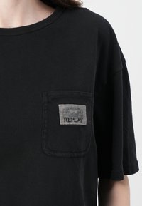 Schwarzes T-Shirt aus weicher Baumwolle, mit einer Brusttasche und einem grauen rechteckigen Etikett, auf dem "REPLAY" steht. Kurze Ärmel und runder Ausschnitt.