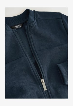 Marineblauer Reißverschlussjacke aus strukturiertem Stoff, mit geripptem Kragen und silbernem Reißverschluss, ausgestattet mit einer seitlichen Tasche und einem minimalistischen Design.