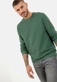 Green ribbed knit sweater with a round neckline and cuffed sleeves, featuring a small embroidered logo on the left arm, paired with gray jeans.