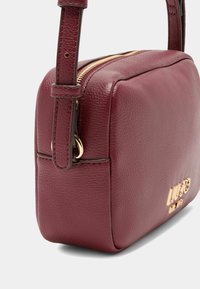 Burgundské kožené crossbody taška s obdélníkovým tvarem, zlatými kovovými prvky a texturovaným povrchem. Obsahuje zipový uzávěr a nastavitelný popruh.