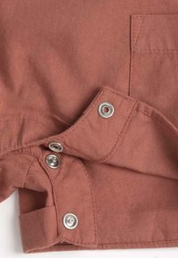 Tissu couleur rouille avec des boutons-pression argentés et une poche. Le matériau a une texture lisse avec des détails de couture visibles.