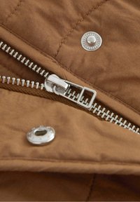 Gros plan sur une fermeture éclair en argent et des boutons-pression sur un tissu matelassé marron, montrant les détails de la couture et la texture.