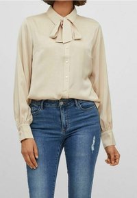 Femme portant un chemisier beige à manches longues avec un col noué et un jean bleu taille haute avec une légère déchirure sur la cuisse.