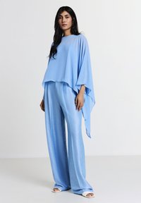 Top azzurro chiaro, morbido e svasato in tessuto trasparente abbinato a pantaloni blu lucidi e larghi. L'outfit presenta un design fluido e una texture a strati.