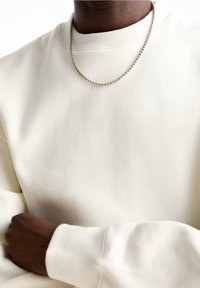 Personne à la peau foncée portant un sweat crème et un collier en argent, le bras croisé sur la poitrine.