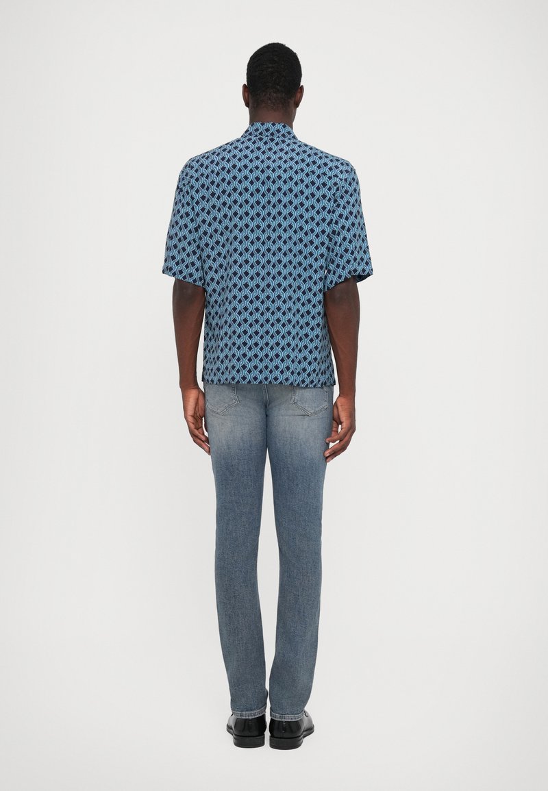 Hombre de espaldas vistiendo una camisa de manga corta azul con patrón, jeans rectos deslavados y zapatos negros, frente a un fondo blanco liso.