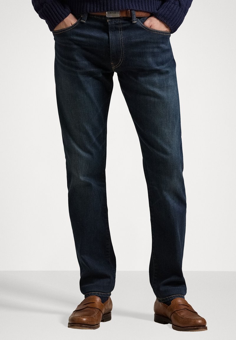 Jeans in denim blu scuro con una vestibilità dritta, caratterizzati da dettagli di cucitura discreti e indossati con scarpe in pelle marrone.