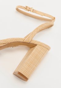 Sandale à talons hauts beige avec un design tissé texturé, une bride de cheville et un talon bloc robuste. Intérieur lisse avec détail en boucle métallique.