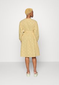 Vestido amarelo com flores, mangas longas, cintura ajustada e uma saia leve. Apresenta pequenos padrões florais em rosa e branco. Vista traseira.