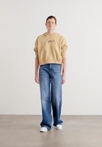 Beige oppkutt hoodie med brodert logo, kombinert med lyseblå vide jeans og hvite sneakers, satt opp mot en enkel bakgrunn.