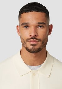 Lichtgekleurde polo met een klassieke kraag, korte mouwen en een knoopsluiting, gedragen over een grijs crewneck-shirt. Glad textuur.