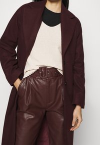 Femme portant un manteau bordeaux foncé, un pull en maille crème, un sous-vêtement noir, et un pantalon en cuir bordeaux taille haute avec une ceinture.