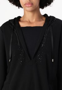 Felpa nera con scollo a V, dotata di decorativi strass neri lungo le cuciture delle spalle e lacci in satin. Materiale in morbido pile.