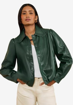 Frau trägt eine dunkelgrüne Kunstlederjacke mit ausgeschnittenen Blumenmustern am Kragen und an den Ärmeln, weißes Oberteil und hellbeige Hose, Hände in den Taschen.