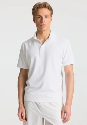 Ung mand med lyst hår iført en ensfarvet hvid kortærmet poloshirt og hvide bukser mod en neutral baggrund.