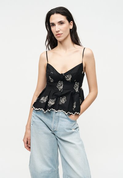 Γυναίκα με μακριά σκουρόχρωμα μαλλιά που φοράει μαύρο αμάνικο τοπ με κεντημένα λουλούδια και κουμπιά, καθώς και ανοιχτό μπλε τζιν παντελόνι με φαρδιά πόδια.