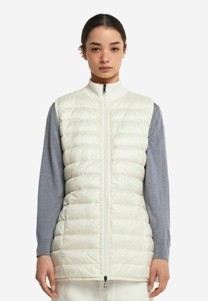 Wattierte Weste in hellem Creme mit gestepptem Muster. Verfügt über einen hohen Kragen und einen Reißverschluss an der Vorderseite. Getragen über einem blauen Langarmshirt.