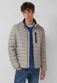 Lichtgrijze, gewatteerde jas met een opstaande kraag en een zwart ritssluiting zak. Voorzien van geribbelde manchetten en een gestructureerd materiaal. Gedragen over een blauwe trui.