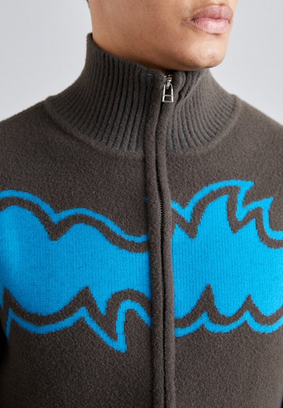 Fleece kapuces džemperis ar augstu ribotu apkakli, brūnu pamatni un zilus viļņveida rakstus ar tumši pelēkiem akcentiem dizaina garumā.